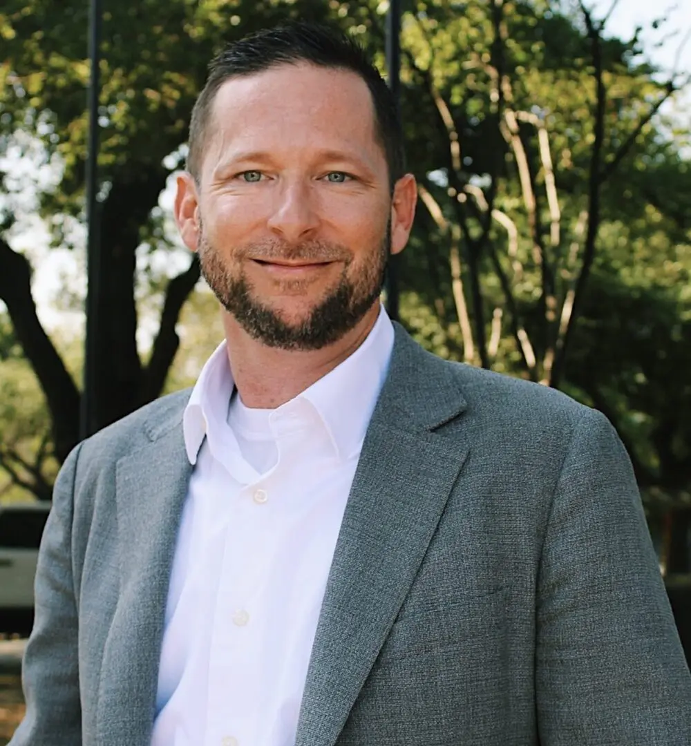 Smiling man in a gray blazer outdoors with trees in the background.